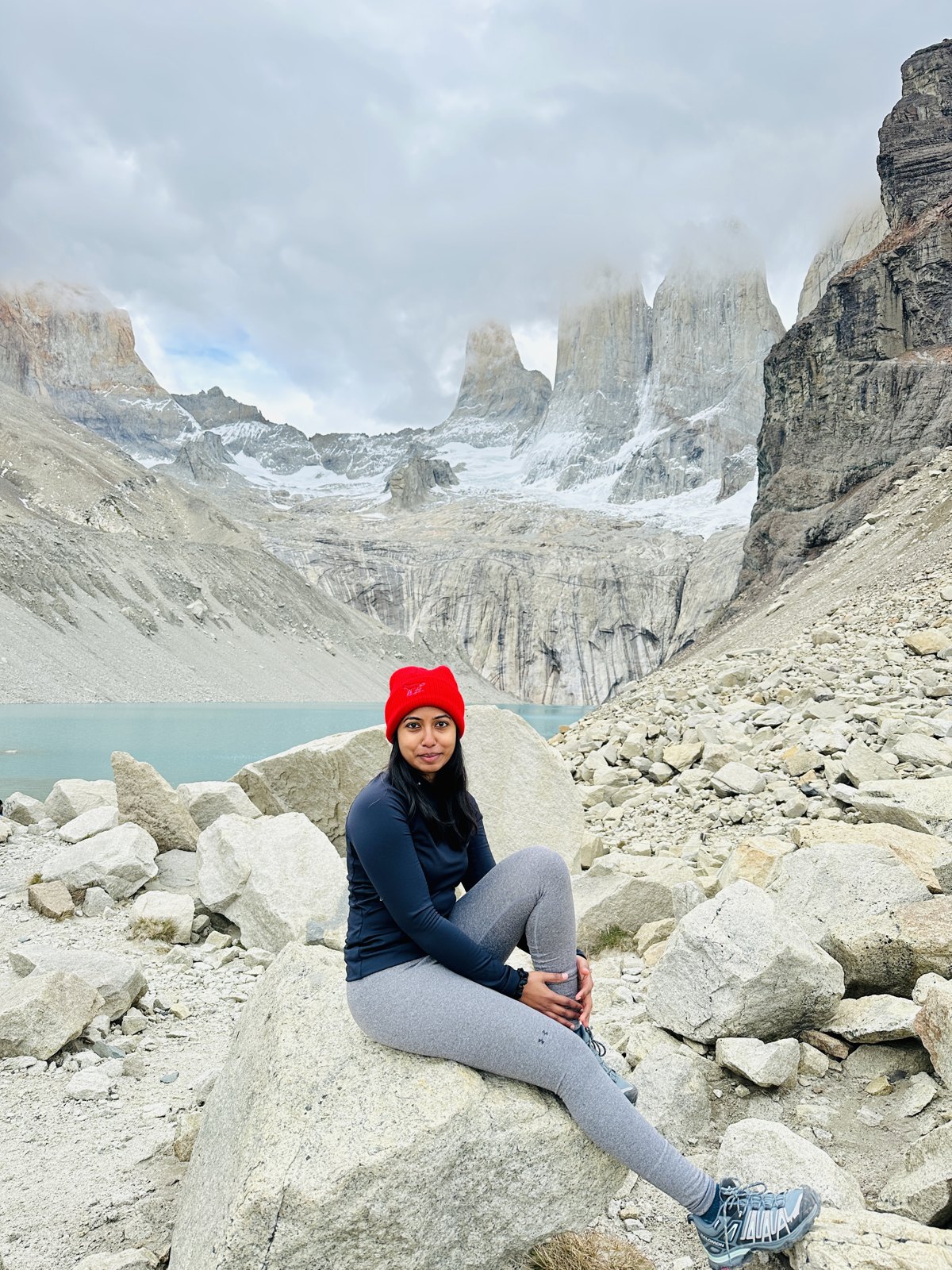 A woman in a red hat sitting on a boulder in front of the three granite towers of Torres del Paine and the pale green lake beneath them
