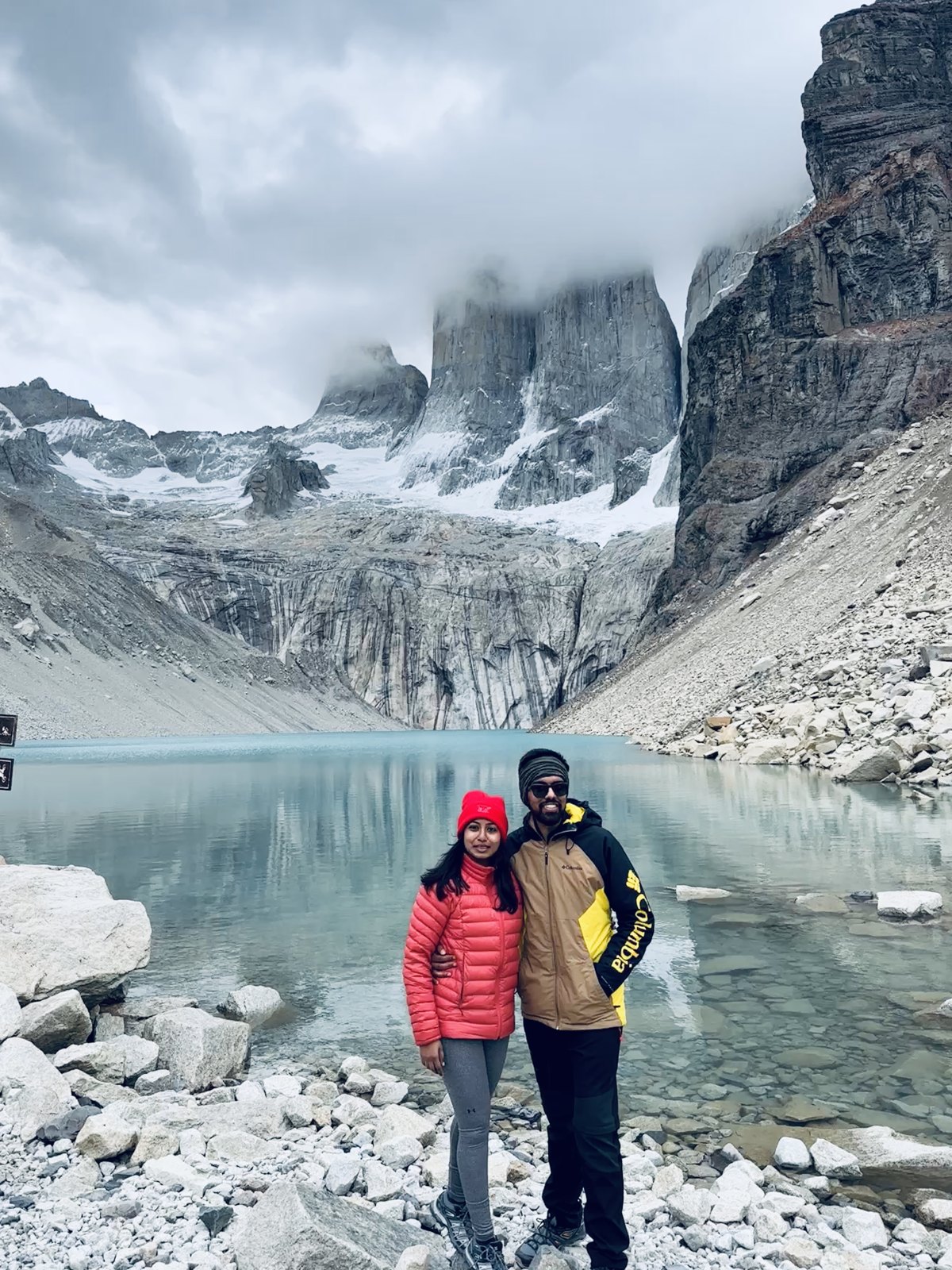 A couple standing on the rocks at the edge of the turquoise lake below the Torres del Paine towers, cloud rolling over the tops