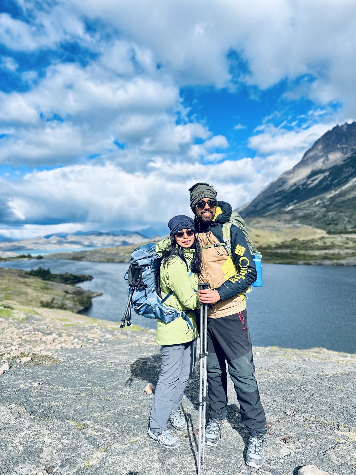 A couple in walking gear with large packs standing together in front of a lake and mountains on the trail