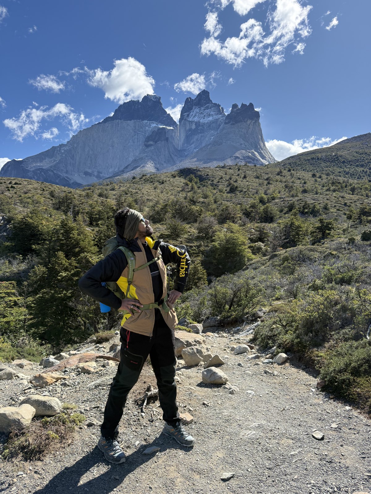 A man with a pack on a stony trail looking up at the dark-capped granite horns of Los Cuernos against a blue sky