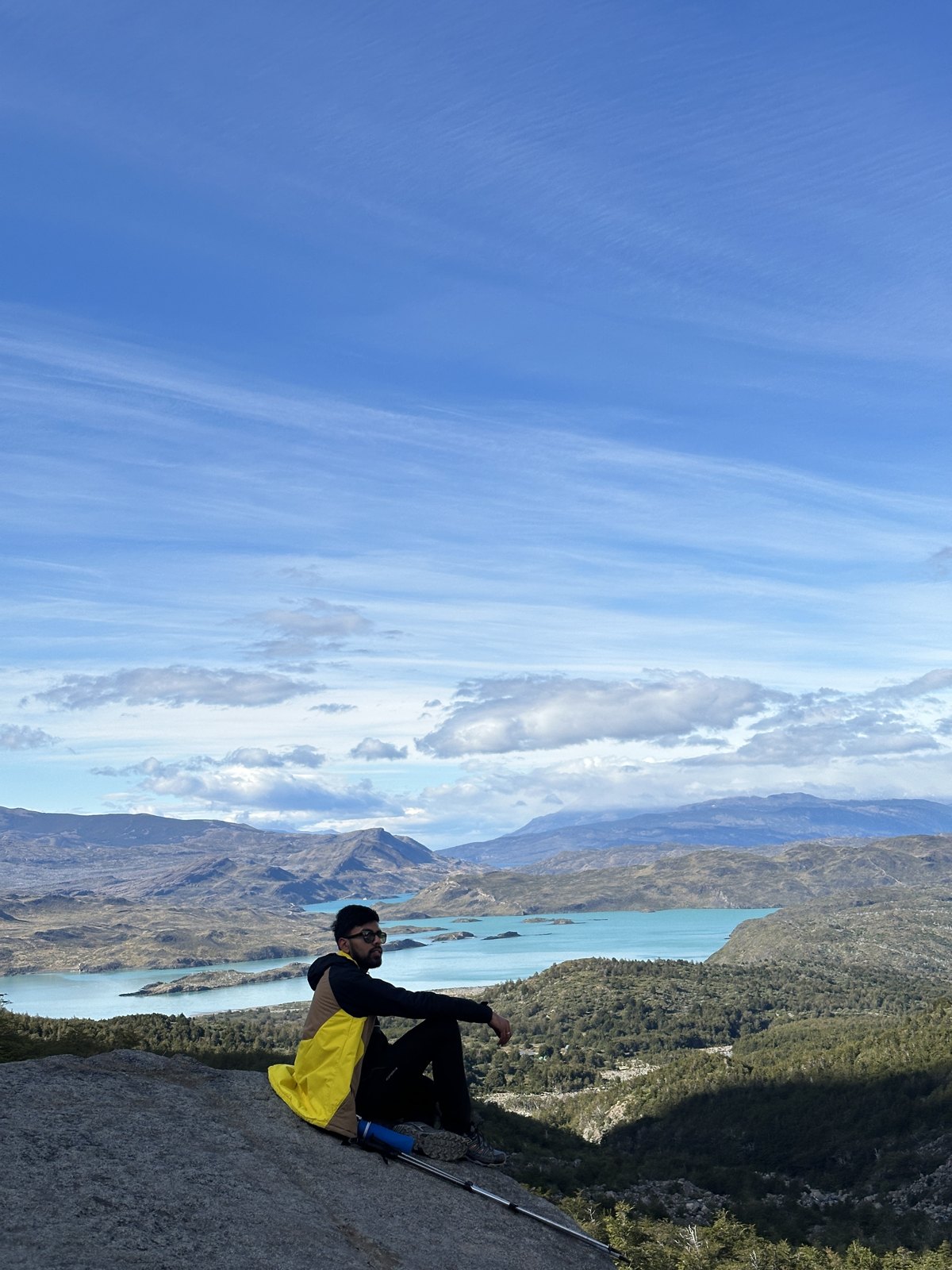 A man sitting on a broad slab of rock at a lookout, with turquoise lakes and the wide valley spread out far below