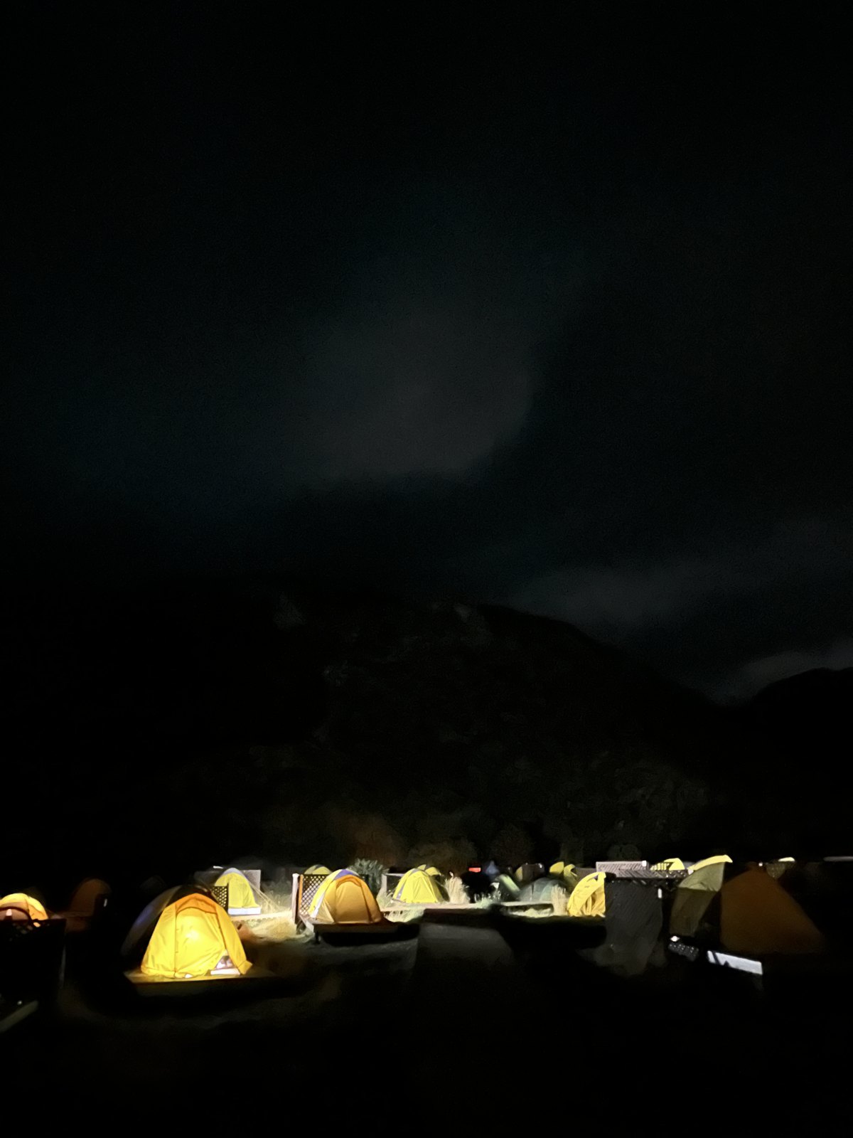 A row of lit tents glowing yellow in the dark at a campsite with a black mountain silhouette behind