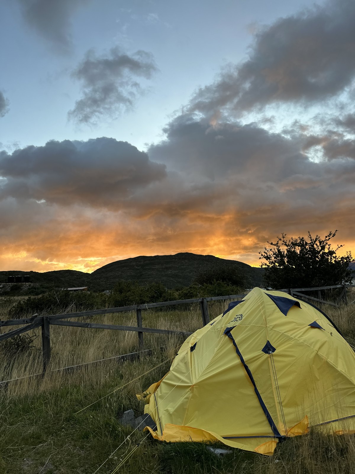 A yellow dome tent pitched in long grass beside a wooden fence, with an orange and grey sunrise over a low hill behind