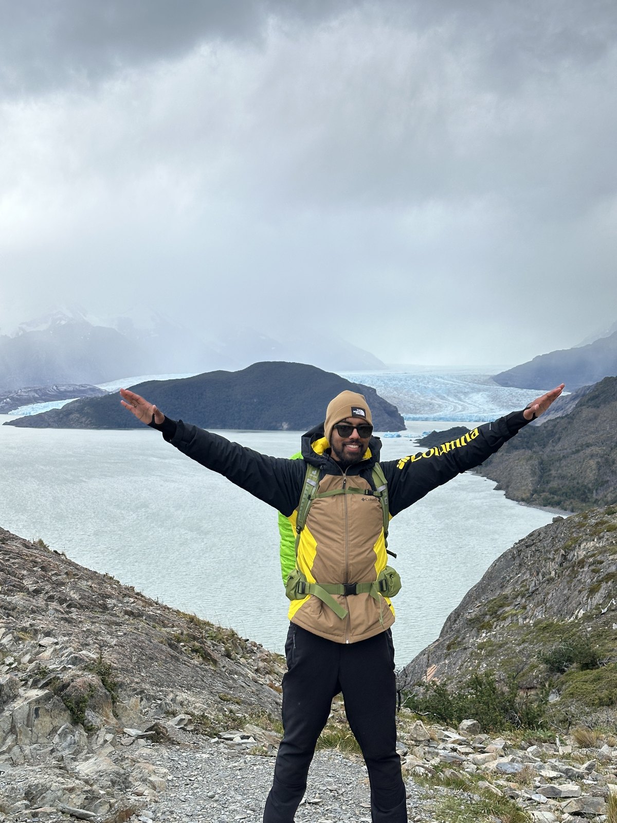A man standing with arms outstretched on a ridge, with the wide grey-blue front of Glaciar Grey across the lake behind him