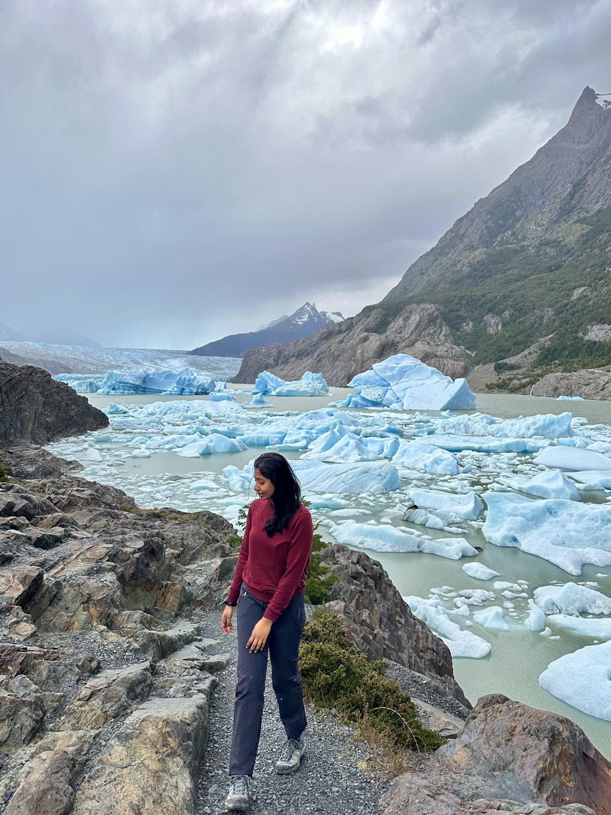 A woman on a rocky shoreline path beside pale blue icebergs floating in the grey water of Lago Grey
