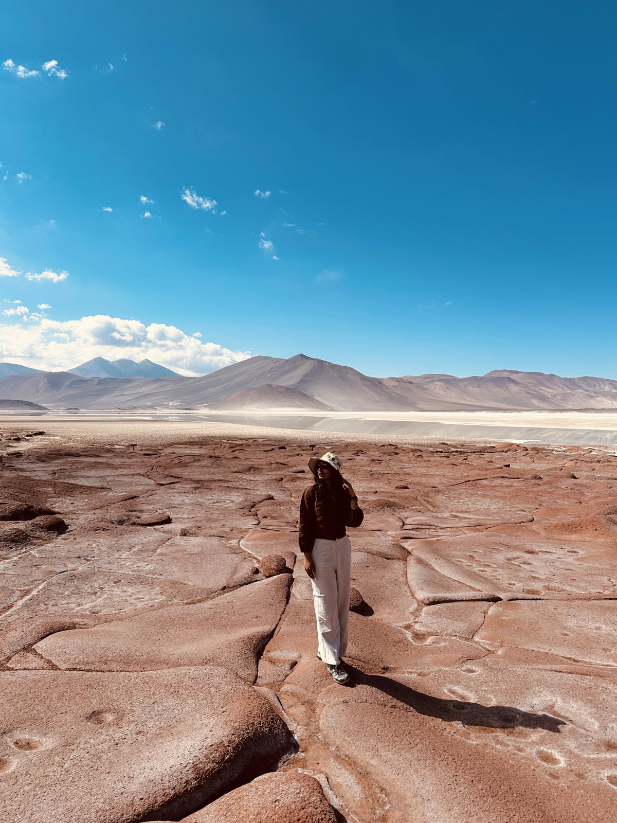 A woman in a hat standing on cracked red rock with a pale salt flat and desert mountains stretching behind her