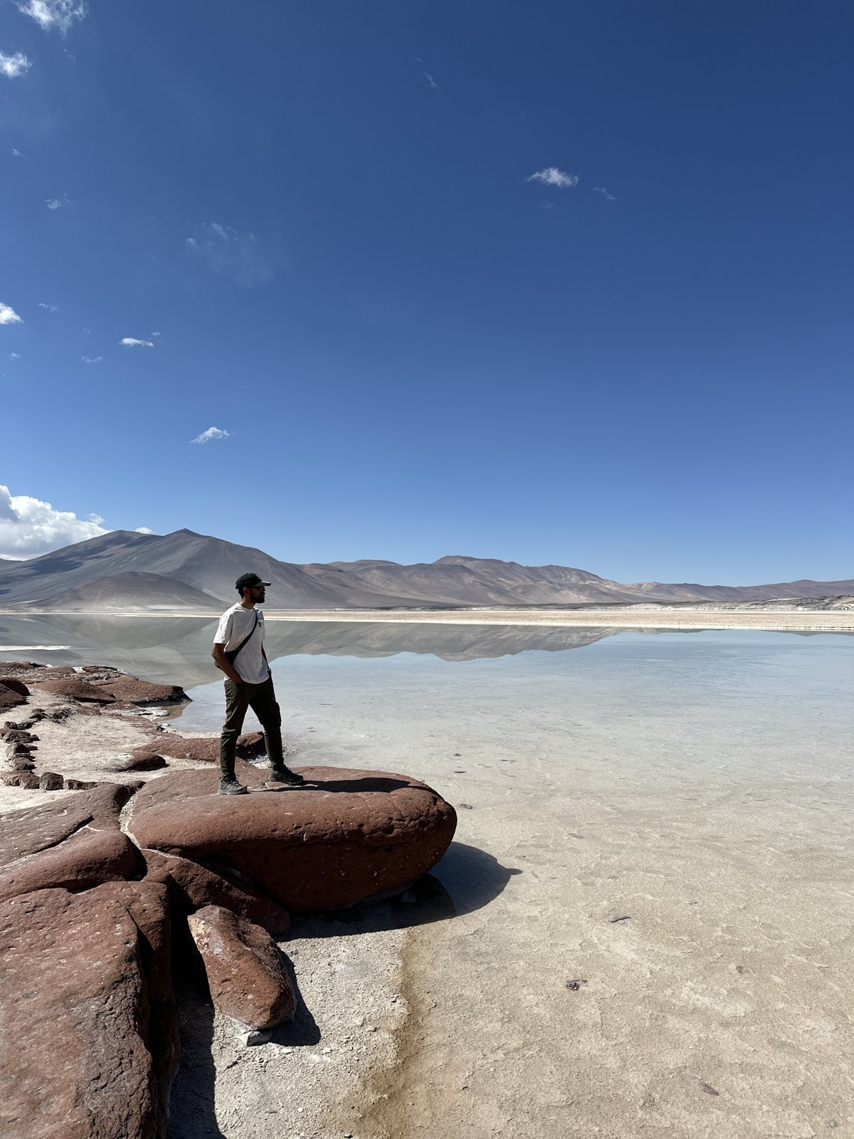A man standing on a red rock outcrop at the edge of a still, mirror-like salt lagoon ringed by desert mountains