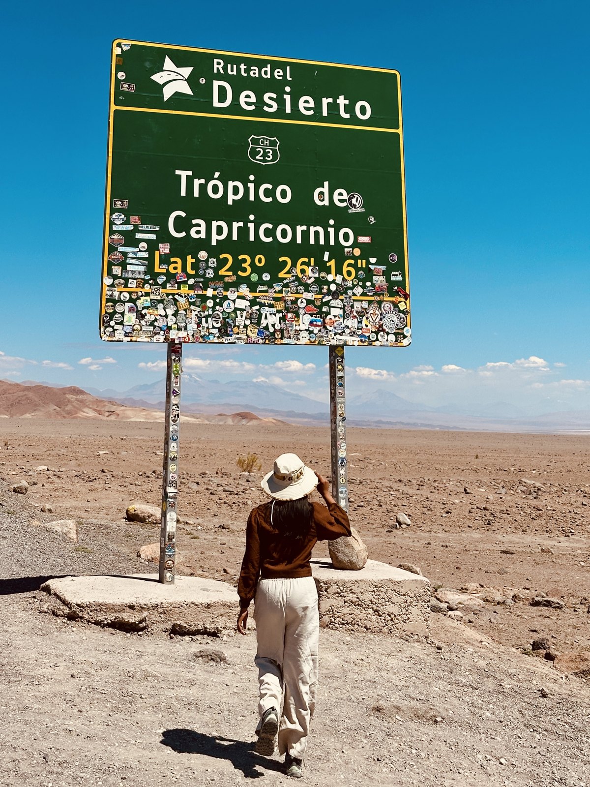 A woman walking towards a large green road sign reading Tropico de Capricornio, covered in stickers, in an empty desert landscape