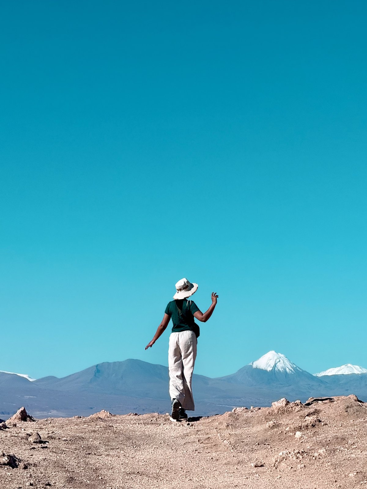 A woman standing on a bare desert ridge with one arm raised mid-movement, snow-capped volcanoes and deep blue sky behind her