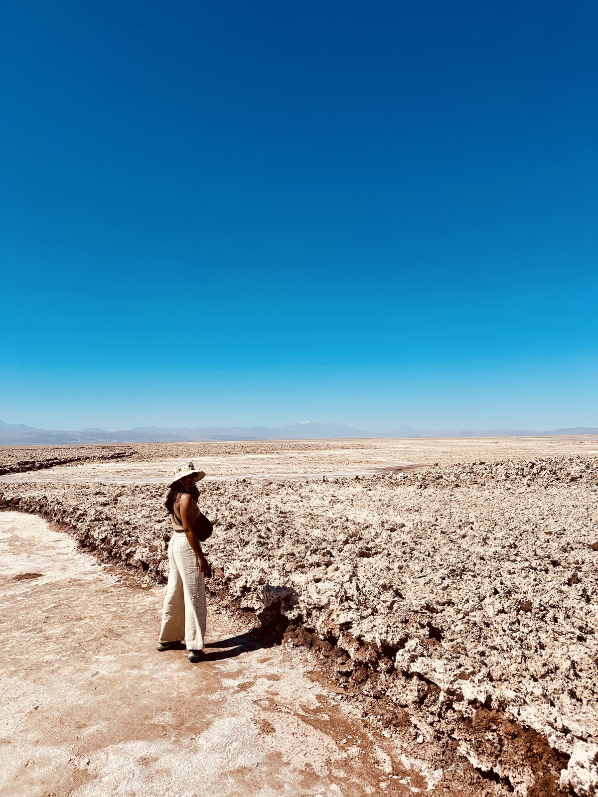 A woman in a wide hat standing beside the rough white and grey crust of the Salar de Atacama, which stretches to the horizon