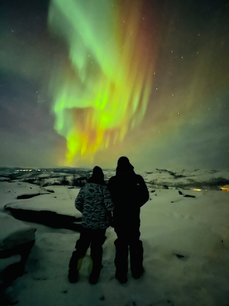 Two people standing in the snow watching a bright green and gold aurora above Alta