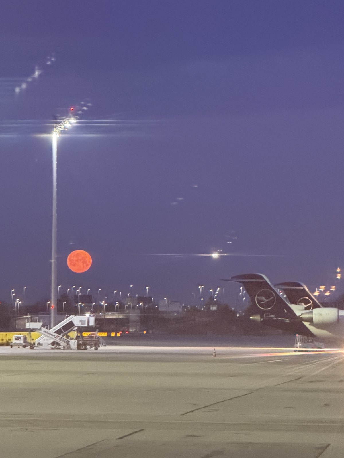 A red moon low over the apron at Munich airport before dawn, with an aircraft tail and ground lights