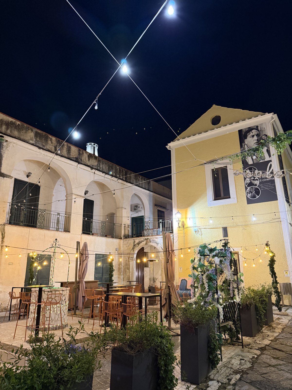A small Italian square at night with festoon lights strung overhead, café tables and a lit yellow building