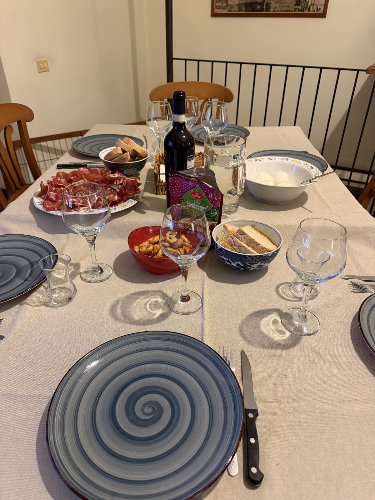 A home dining table laid with plates, wine glasses, a bottle of red wine, a platter of cured meats, taralli and bread