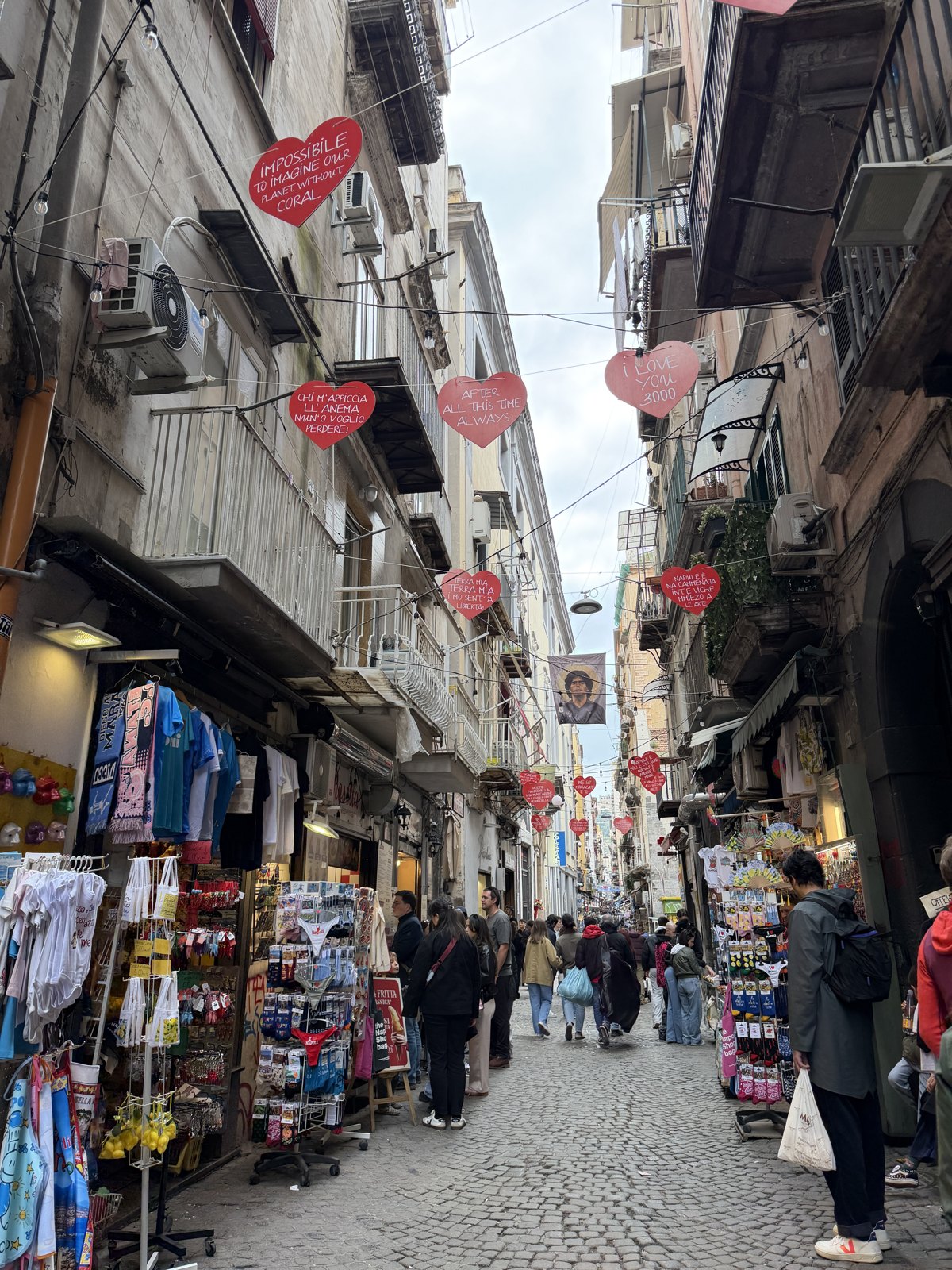 A narrow Naples street lined with market stalls and shops, red heart-shaped signs strung overhead and people walking on the cobbles