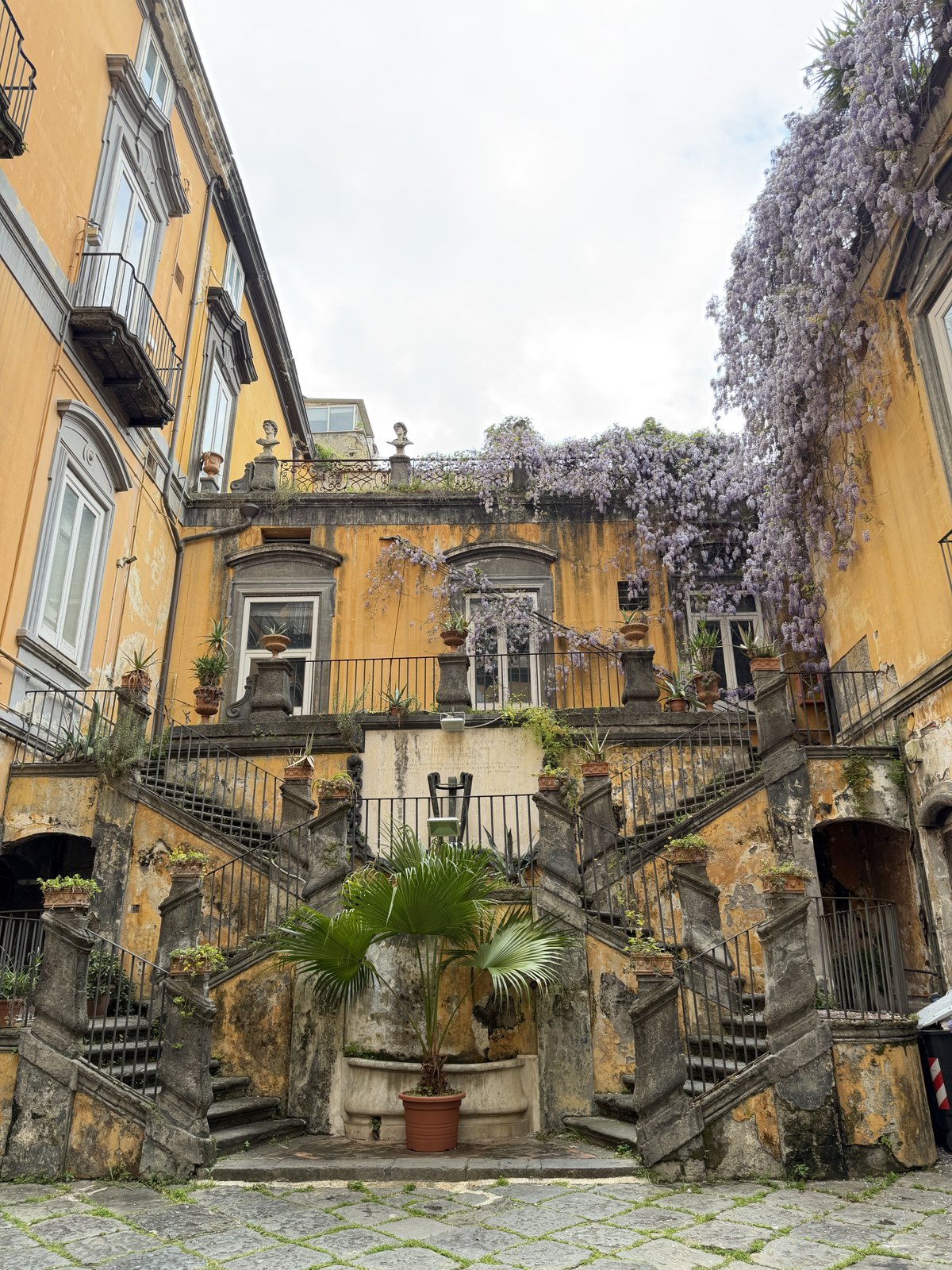 A Naples courtyard with double staircases, ochre walls, potted plants and wisteria hanging over the balustrade