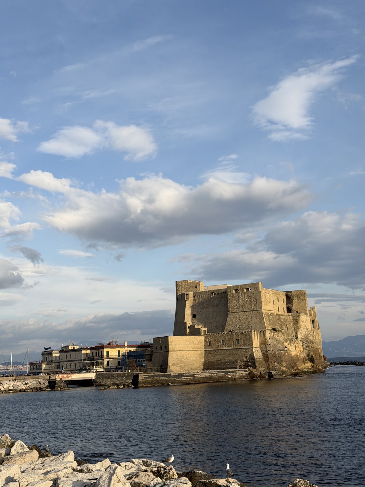 Castel dell Ovo, a large stone fortress on a rocky islet in the sea under a blue sky with clouds