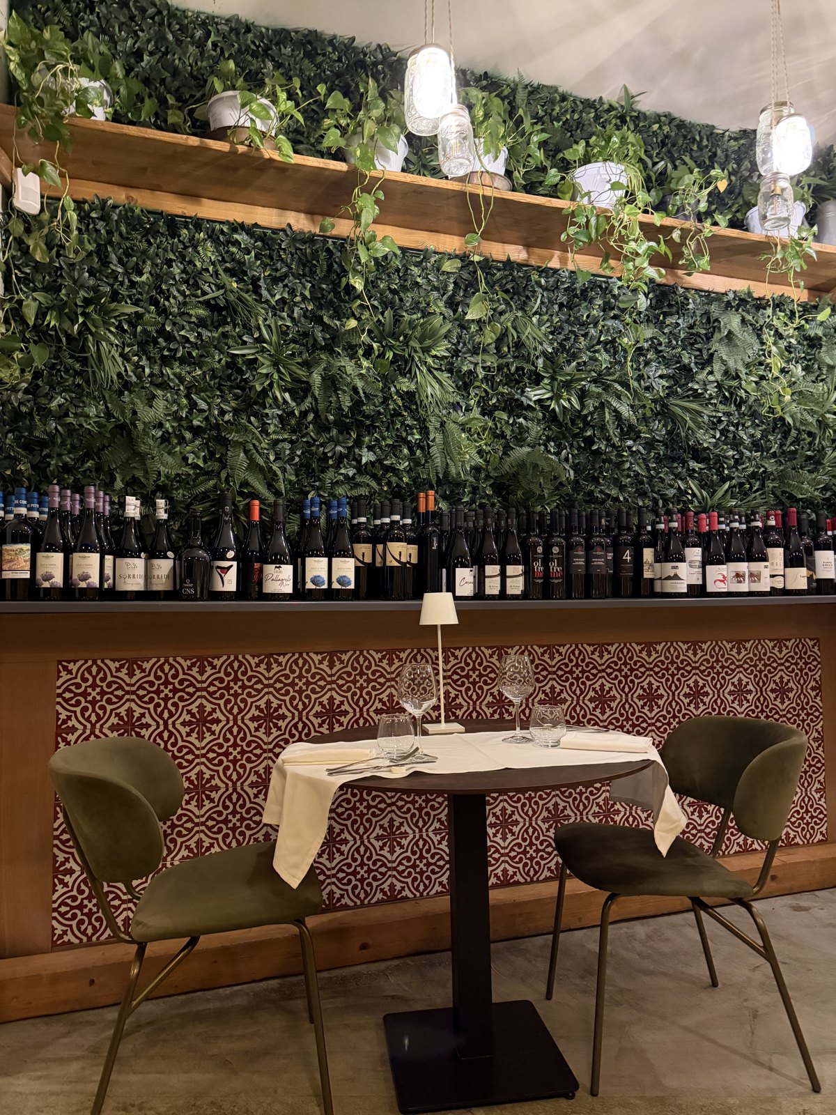 A restaurant table for two set against a wall of greenery with rows of wine bottles behind