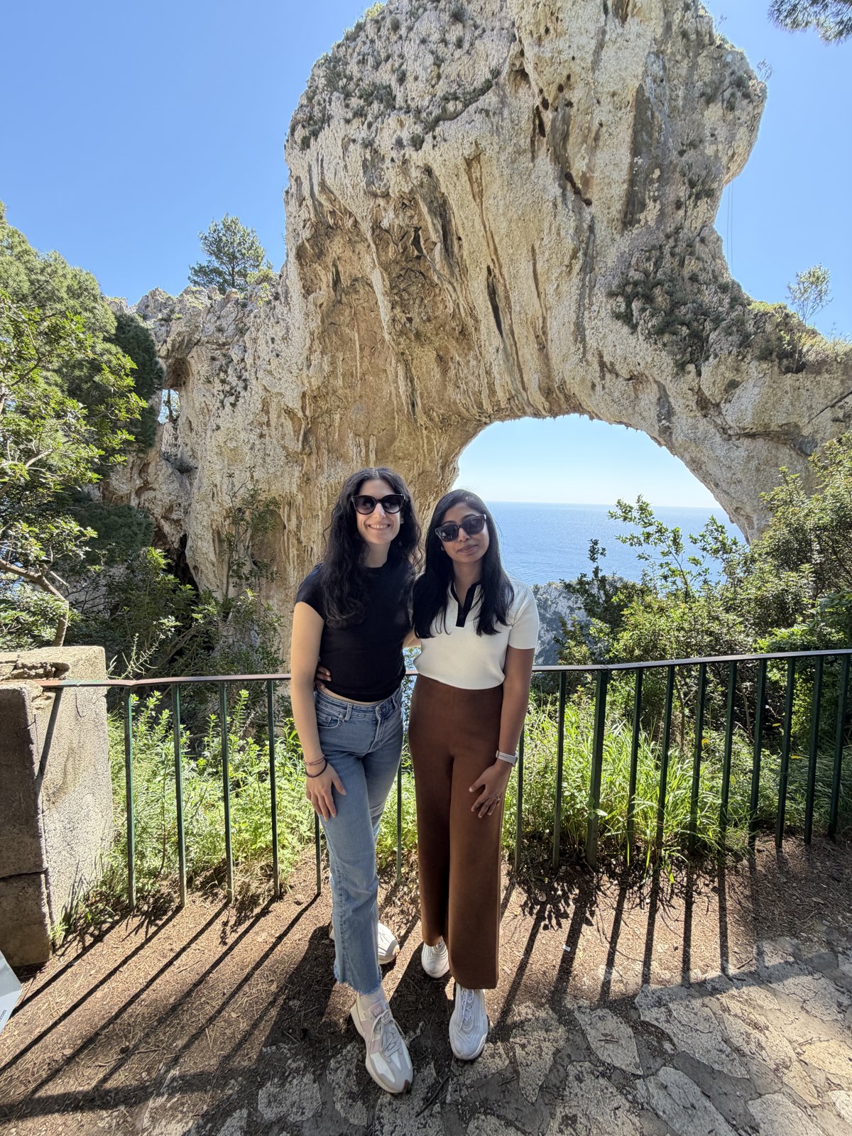 Two women standing under the Arco Naturale, a large natural limestone arch on Capri, with the sea visible through it