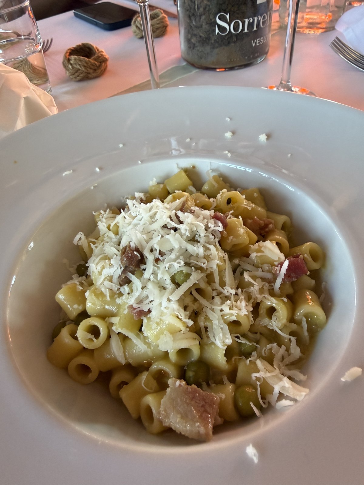 A bowl of short pasta with peas and pieces of cured pork, covered in grated cheese