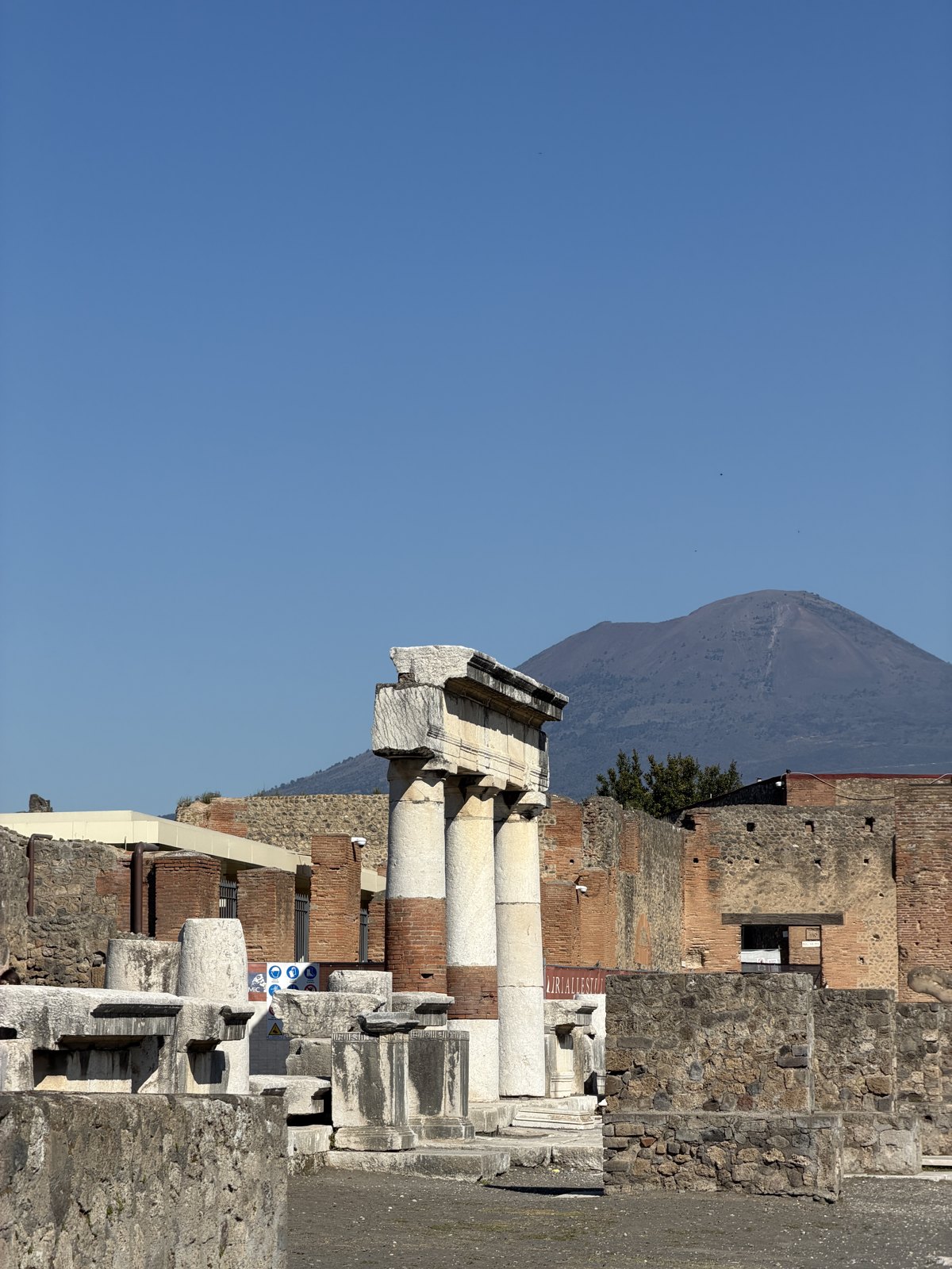 Ruined columns and walls of the Pompeii forum with the cone of Mount Vesuvius rising behind under a clear sky