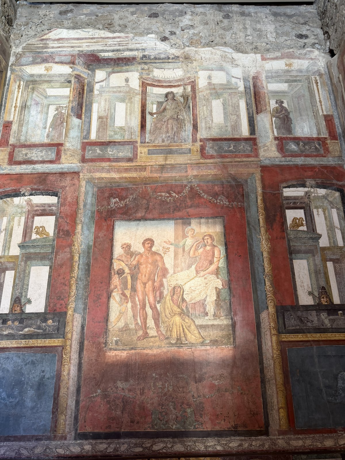A large Pompeian wall painting in red and ochre with a mythological scene in a central panel and painted architecture around it