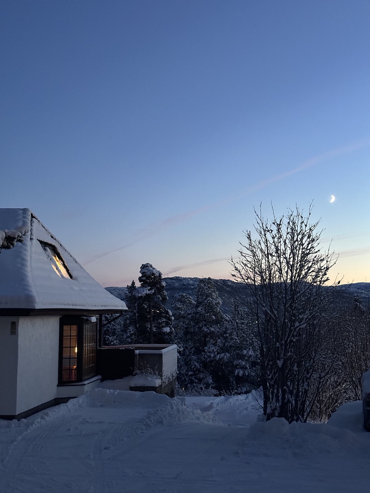 A snow-covered cabin with a lit window at dusk, with pink light on the horizon and a moon above