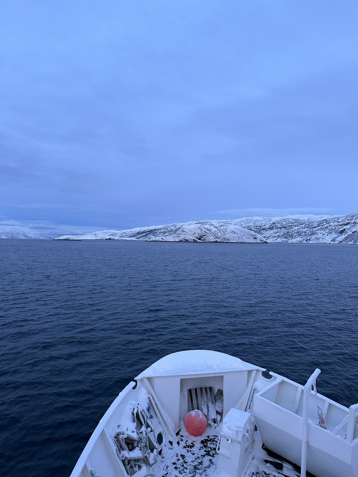 The bow of a boat with snow on the deck, heading across a fjord towards snow-covered hills under a pale sky