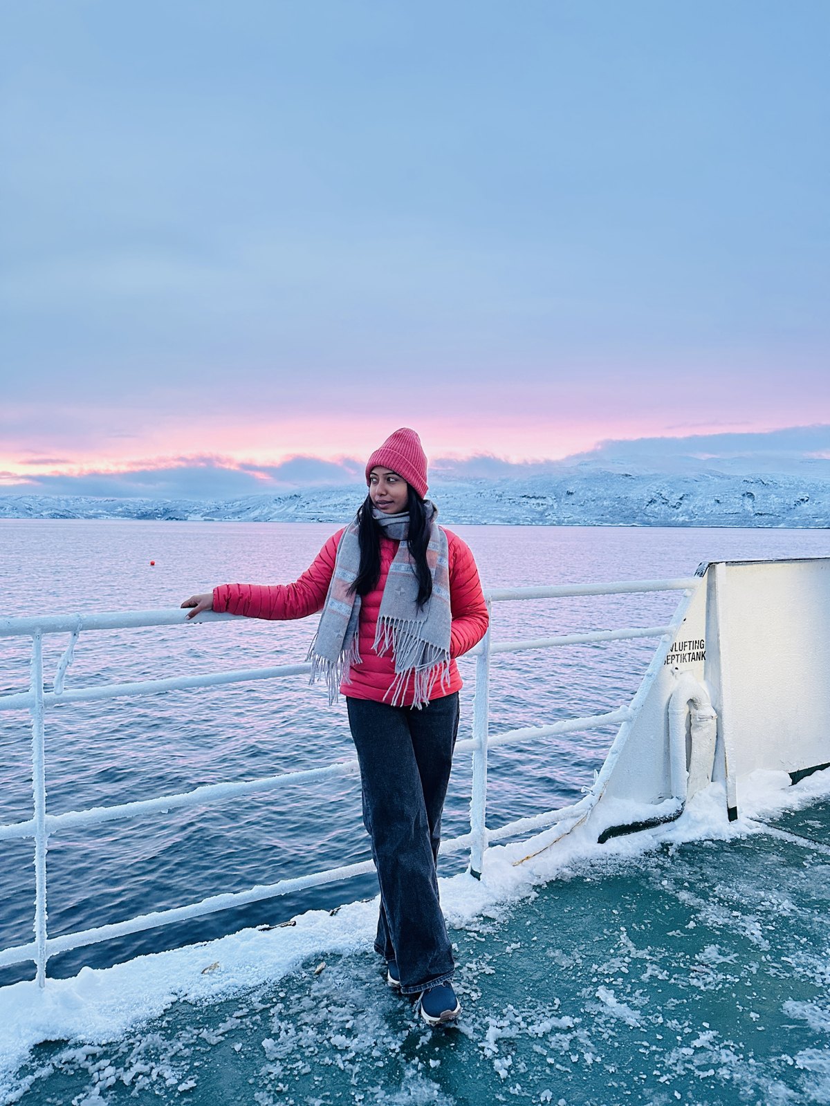 A woman in a red jacket and pink hat standing at the rail of a boat with a pink and blue sunrise sky over snowy hills