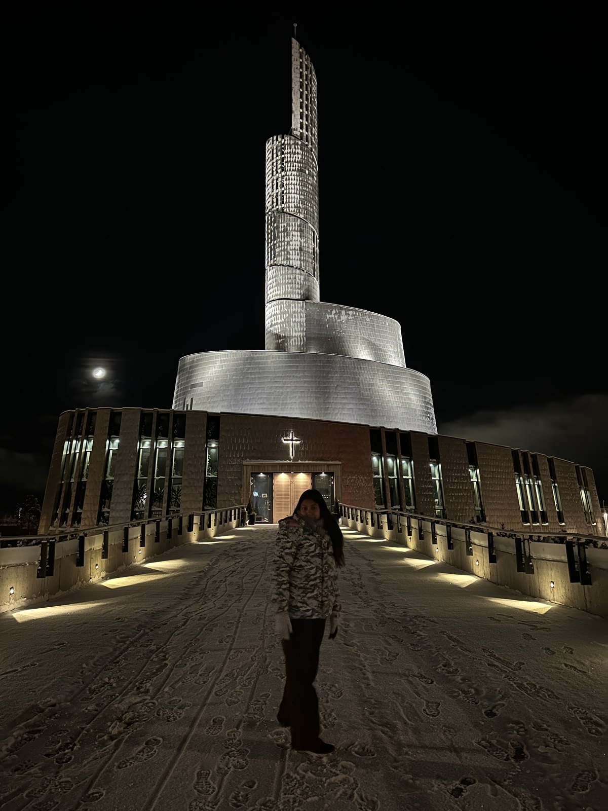 A woman standing in the snow in front of the Northern Lights Cathedral in Alta, its titanium spiral tower lit at night