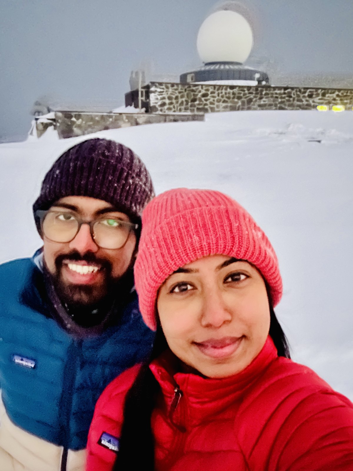 A couple in winter hats taking a selfie in deep snow, with the North Cape hall and its dome behind them