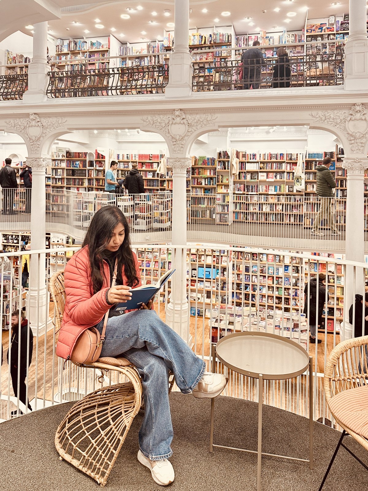 A woman sitting in a wicker chair reading a book on a balcony inside the white multi-storey atrium of the Carturesti Carusel bookshop