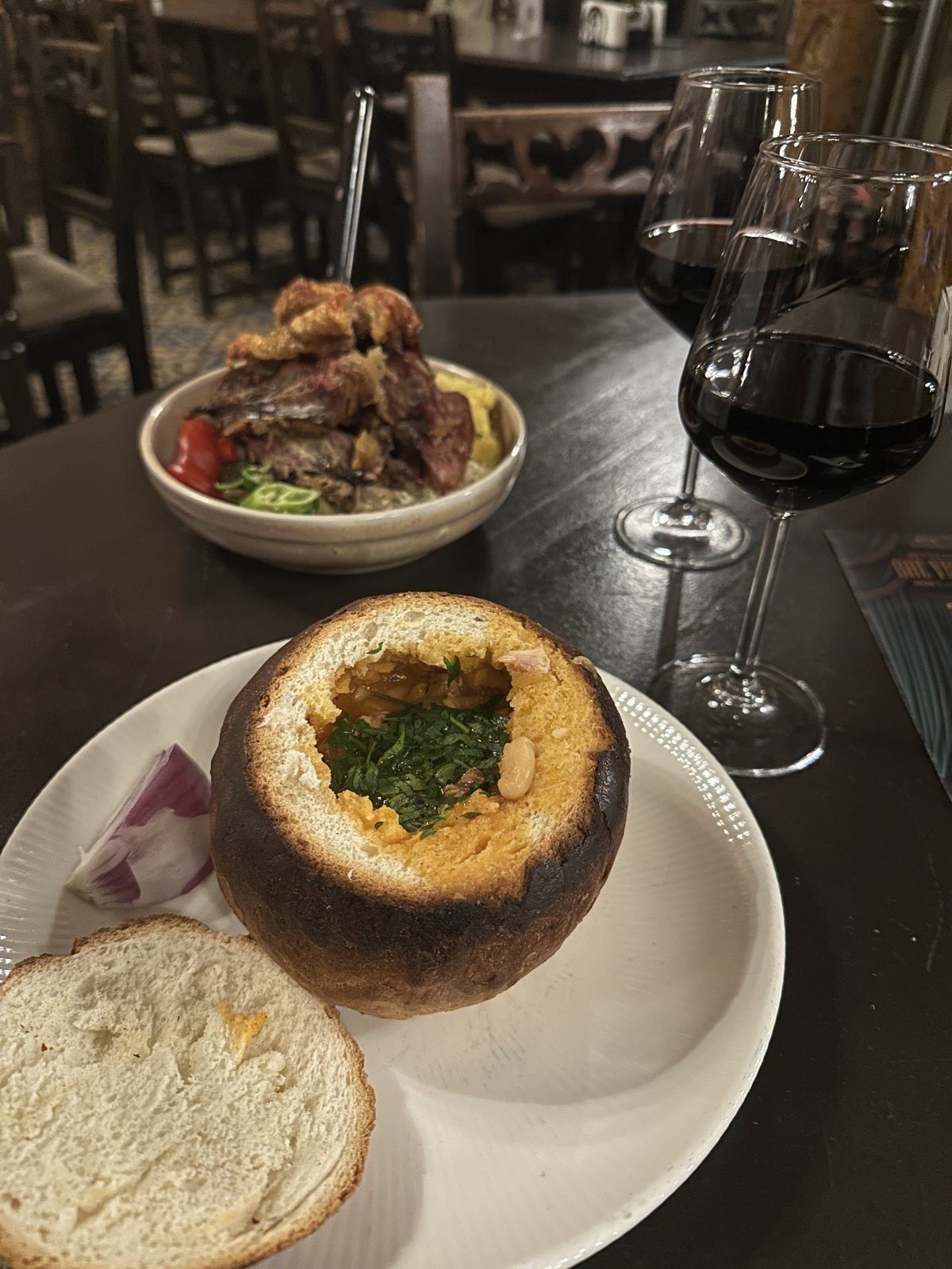 A sour soup served inside a hollowed-out round loaf of bread, with a plate of grilled meats and two glasses of red wine behind it