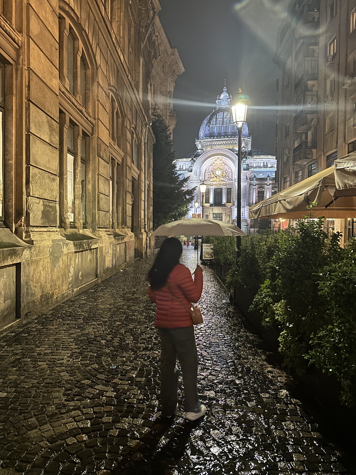 A woman with an umbrella walking down a wet cobbled street at night, with the illuminated dome of the CEC Palace at the end of it
