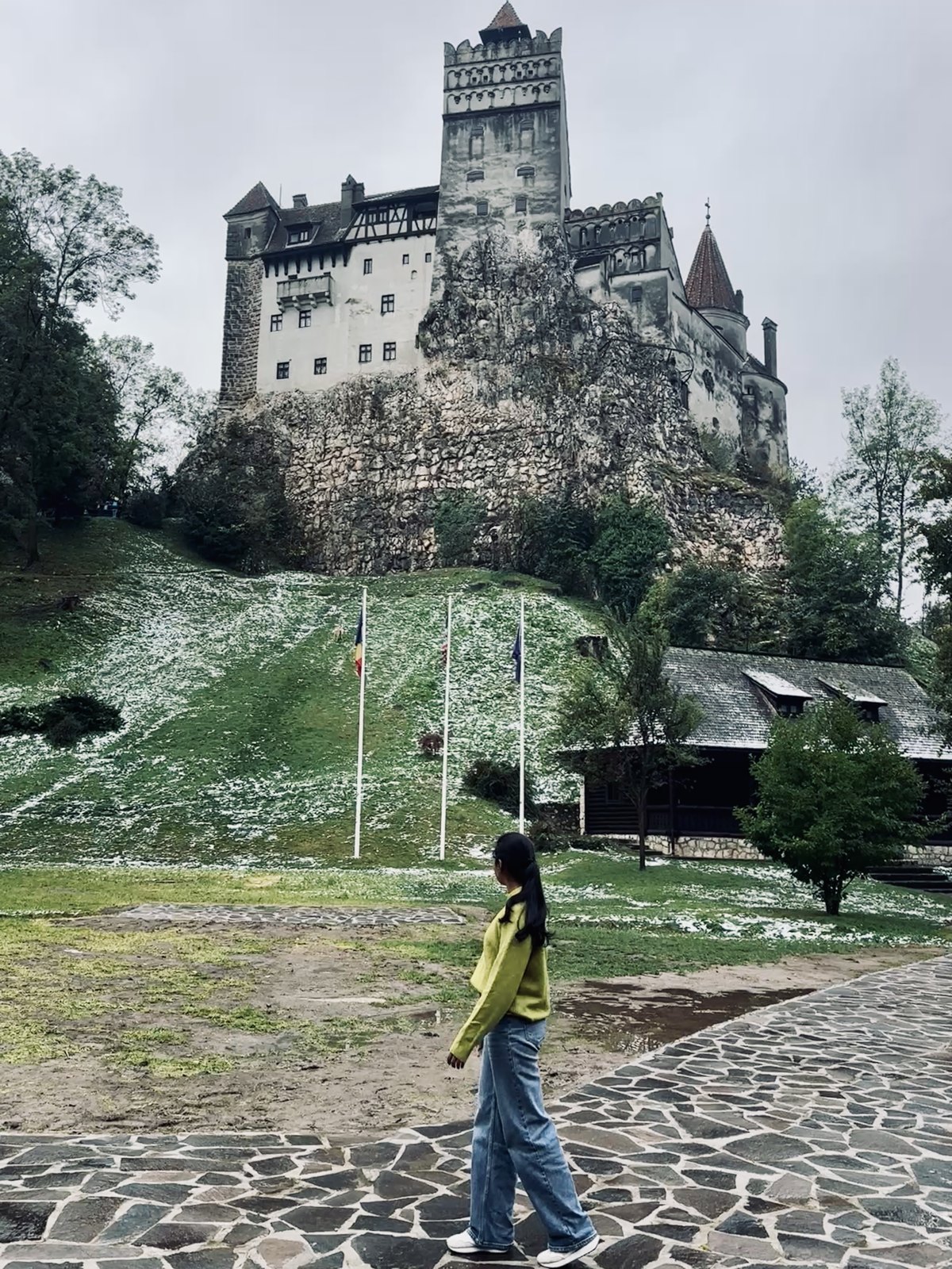 Bran Castle on its rock above a green slope dusted with white hail, with a woman in a yellow jacket walking past in the foreground