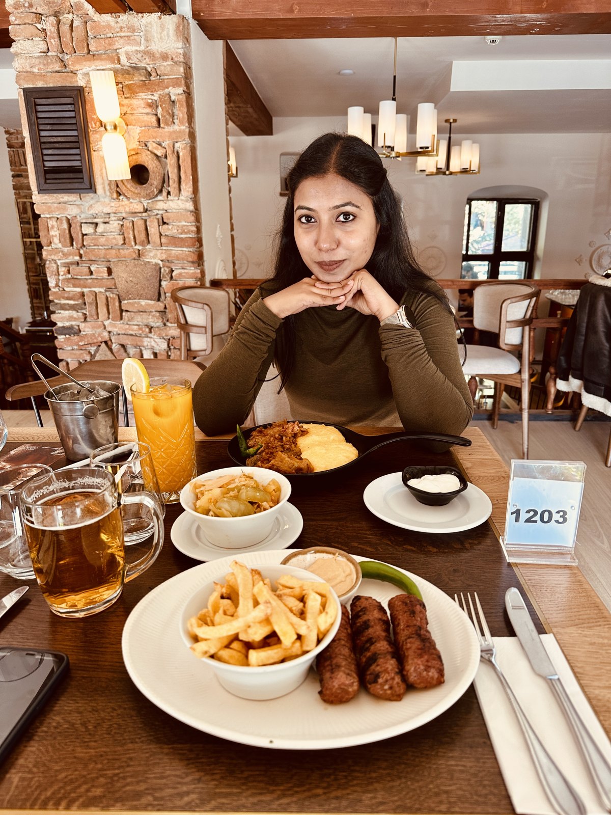 A woman at a restaurant table with a plate of mici sausages and chips, a beer and other dishes in front of her