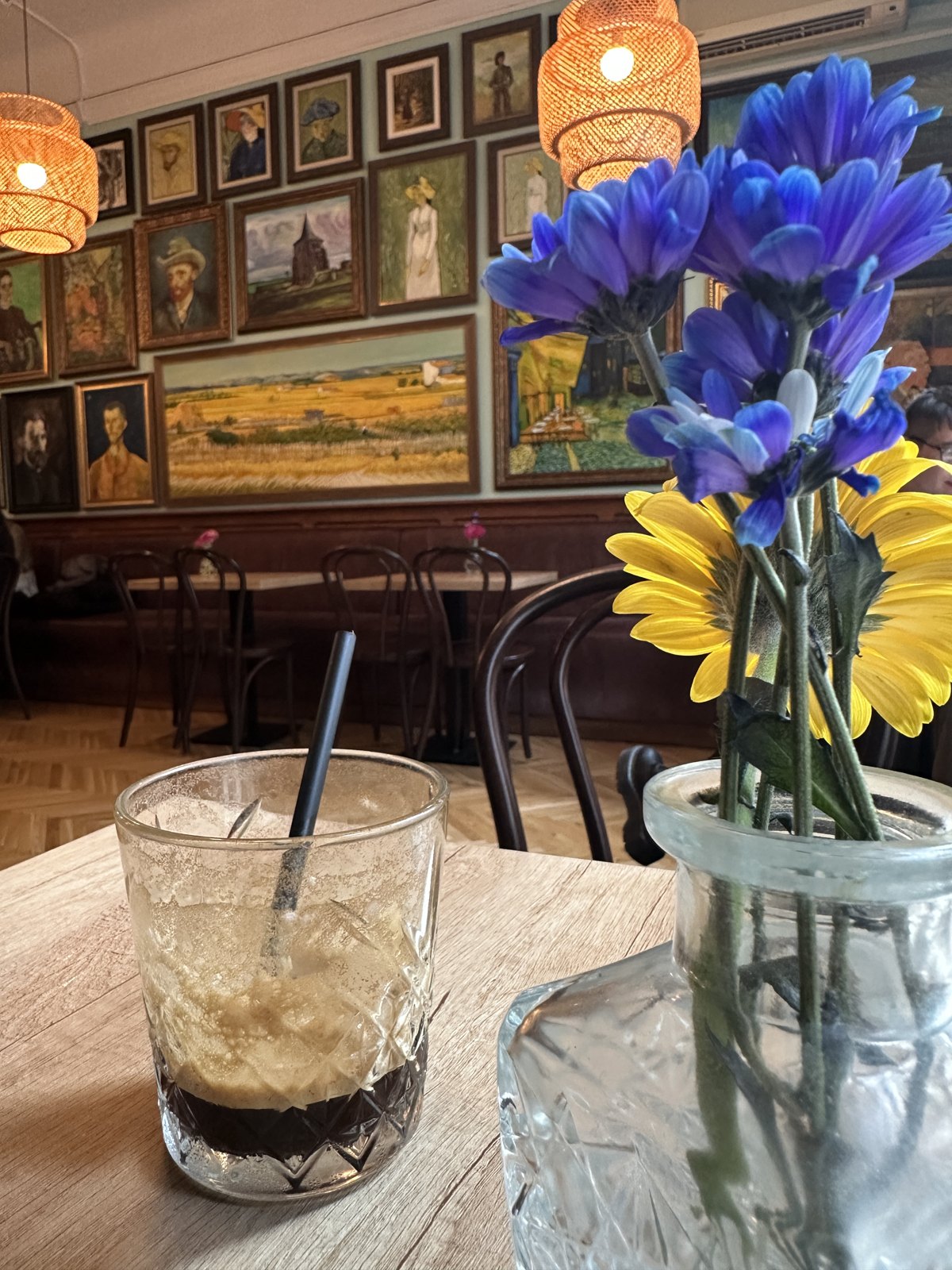 A glass of iced coffee on a cafe table beside a jar of blue and yellow flowers, with a wall densely hung with paintings behind