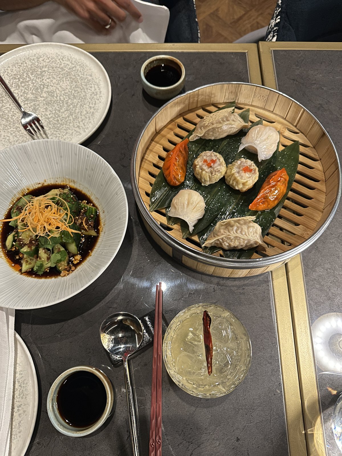 An overhead view of a bamboo steamer of assorted dim sum, a bowl of smashed cucumber salad, and a cocktail