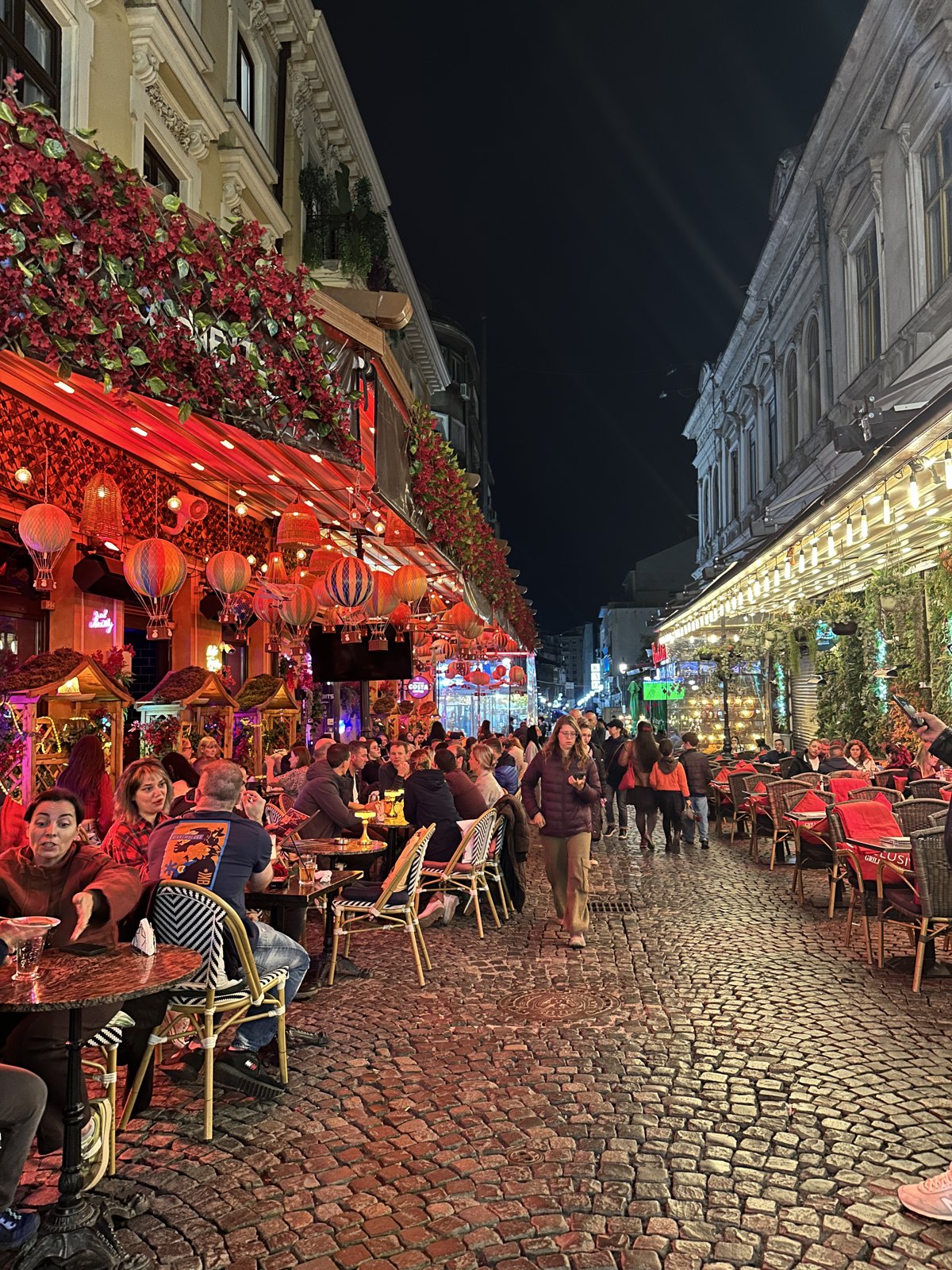 A busy cobbled street in Bucharest old town at night, lined with lit restaurant terraces, red flowers and lanterns