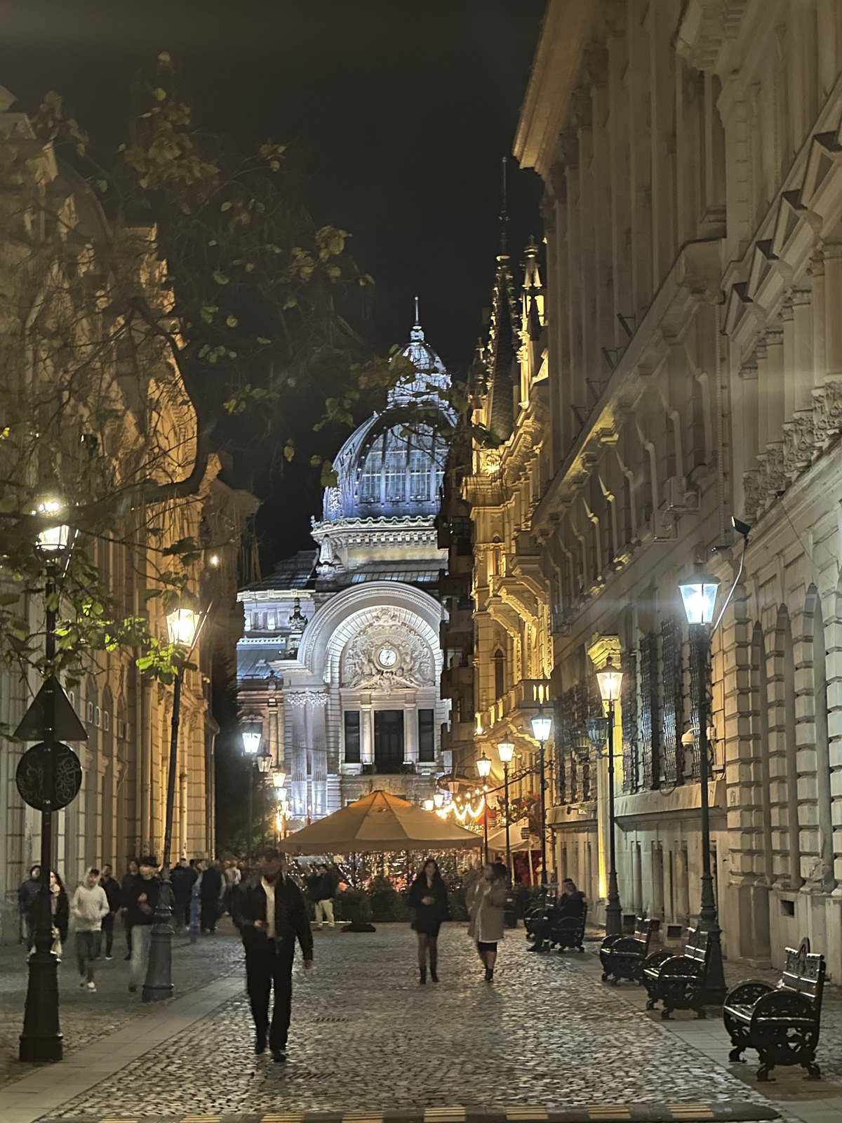 A cobbled street at night with the illuminated glass dome of the CEC Palace at the end of it and people walking