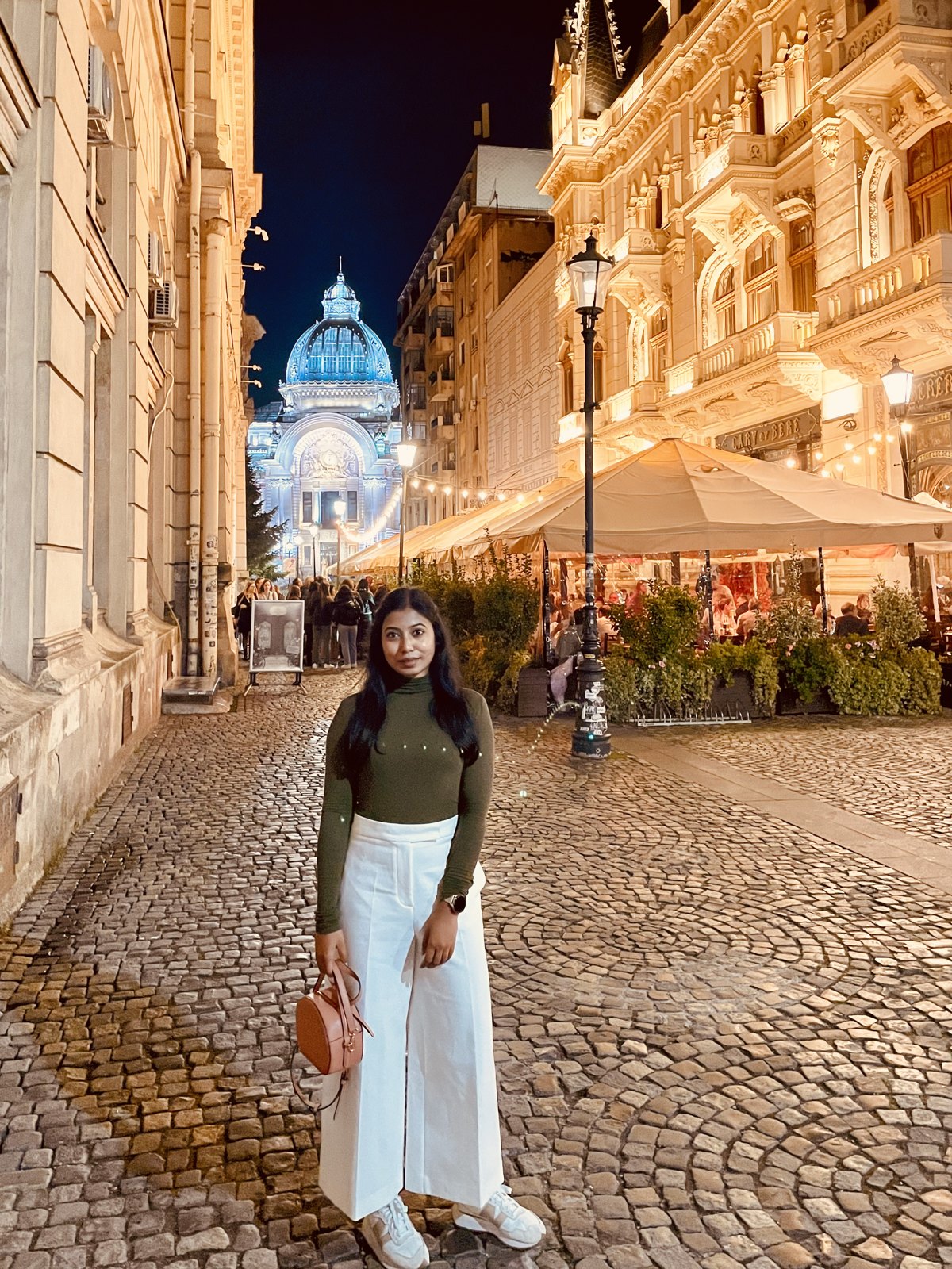 A woman standing on the cobbles of a Bucharest old town street at night, with lit buildings and the CEC Palace dome behind her