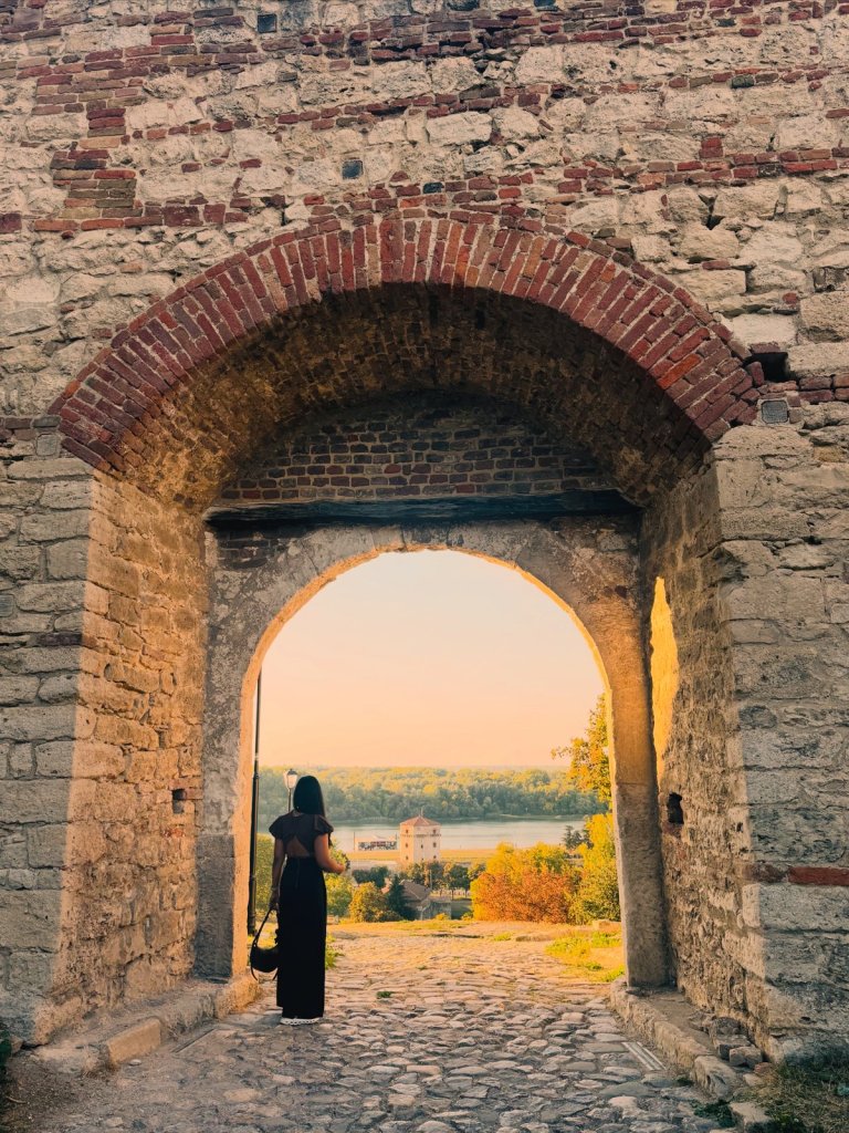 A woman standing in a stone archway at Belgrade Fortress, looking out over the river at sunset