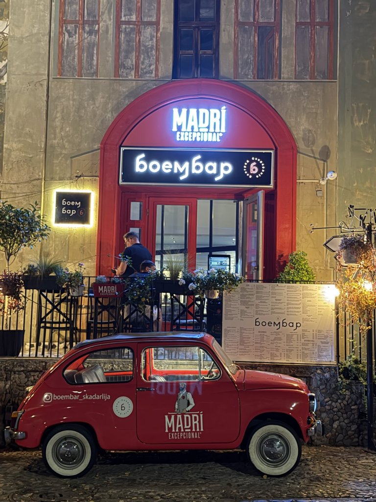 A vintage red car parked outside a bar with a red arched doorway in Skadarlija